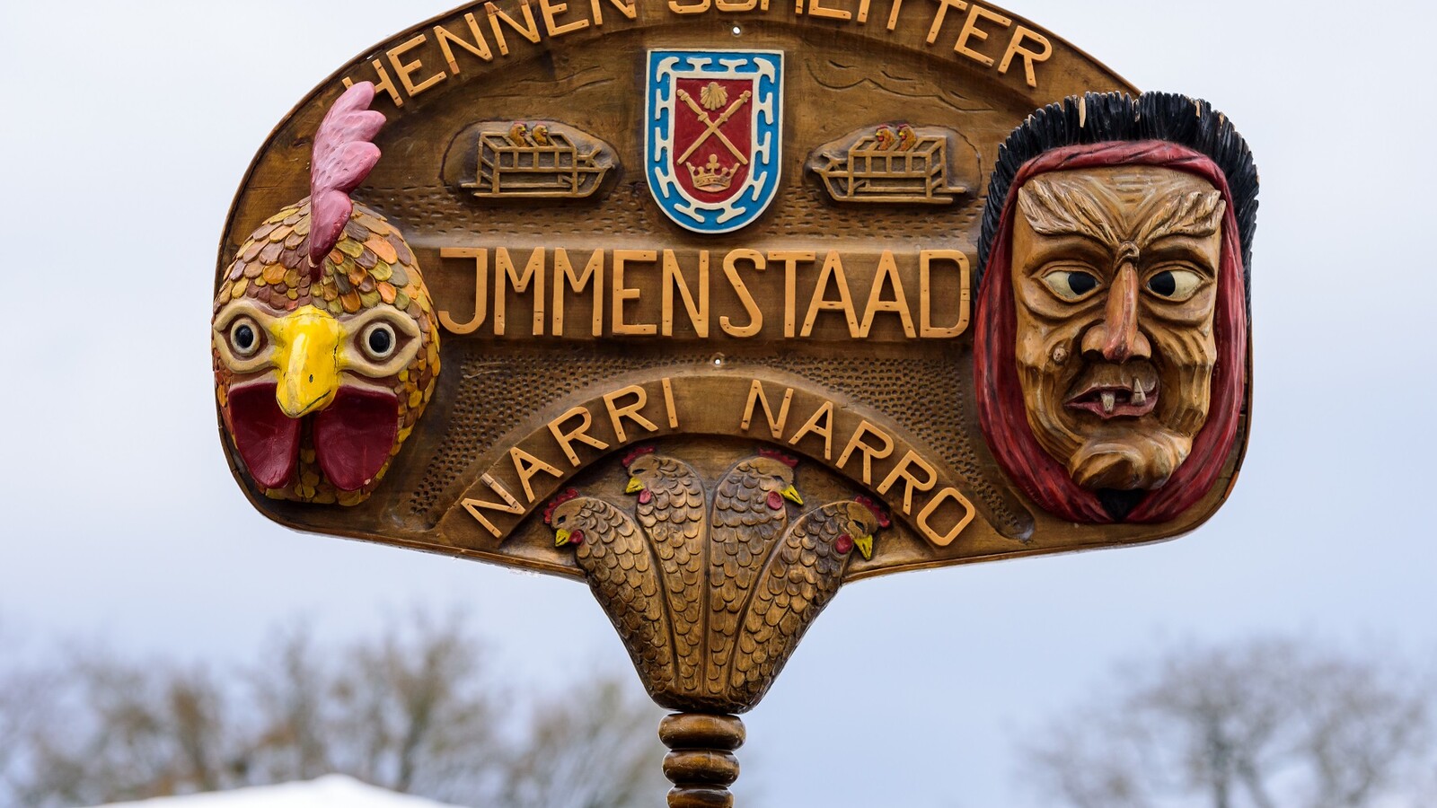Holzschild mit der Aufschrift "Hennenschlitter Immenstaad, Narri Naro" sowie zwei kleinen geschnitzten Masken, die rechts und links abgebildet sind.