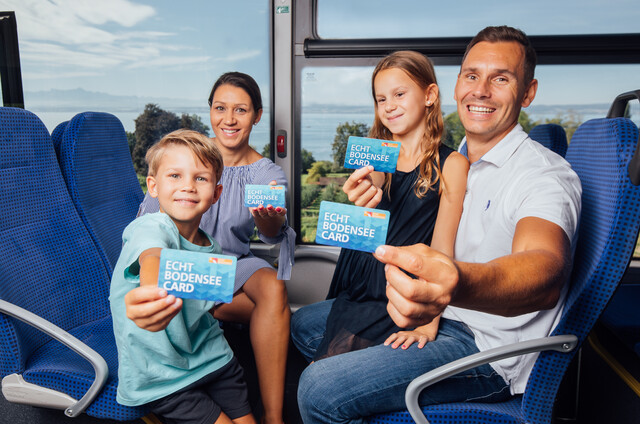 Familie im Bus unterwegs und zeigt die Echt Bodensee Card vor. Blick durch das Busfenster auf den Bodensee.
